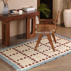Puede incluir: Una alfombra rectangular con un campo crema y un patr&oacute;n repetitivo de diamantes granates. La alfombra tiene un borde azul claro y flecos. Una silla y un escritorio de madera est&aacute;n en el fondo.