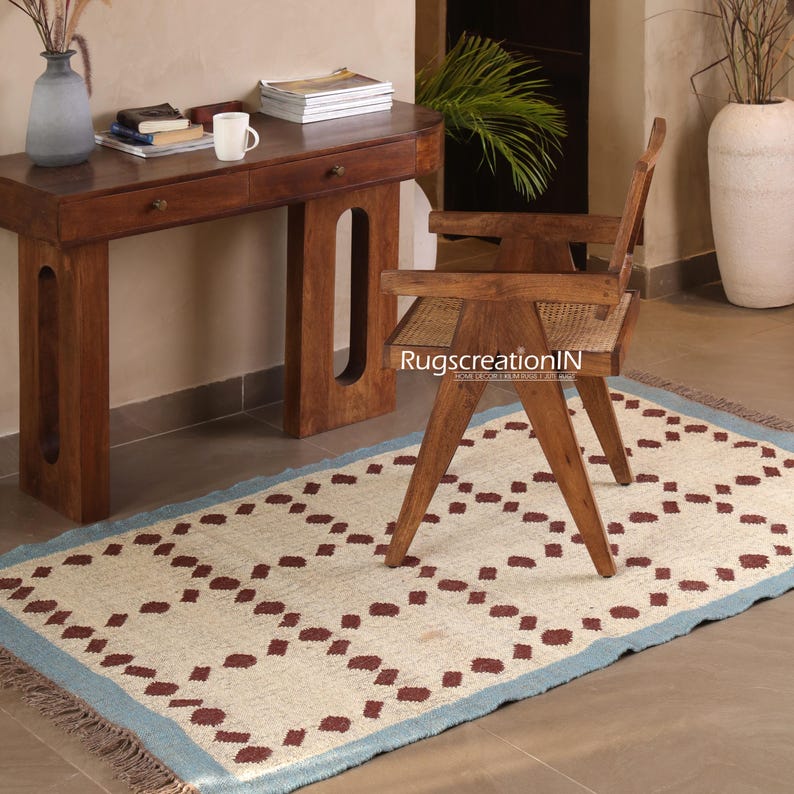 Puede incluir: Una alfombra rectangular con un campo crema y un patr&oacute;n repetitivo de diamantes granates. La alfombra tiene un borde azul claro y flecos. Una silla y un escritorio de madera est&aacute;n en el fondo.