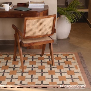 Op de afbeelding: Een houten stoel met een geweven rugleuning en zitting staat op een patroonkleed. Het kleed heeft een geometrisch ontwerp met bruine vierkanten, zwarte lijnen en roze accenten. De stoel staat voor een houten bureau.