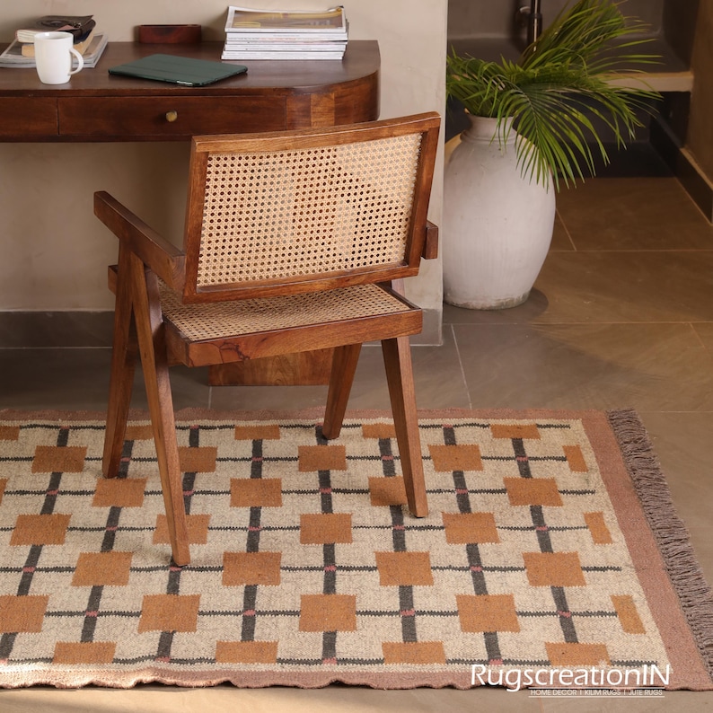 Op de afbeelding: Een houten stoel met een geweven rugleuning en zitting staat op een patroonkleed. Het kleed heeft een geometrisch ontwerp met bruine vierkanten, zwarte lijnen en roze accenten. De stoel staat voor een houten bureau.