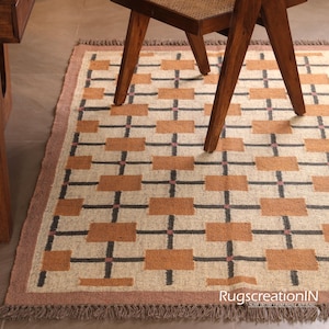 Op de afbeelding: Een rechthoekig vloerkleed met een geometrisch patroon van bruine rechthoeken en zwarte lijnen op een beige achtergrond. Het vloerkleed heeft een bruine rand en franjes. Een houten stoelpot is zichtbaar.