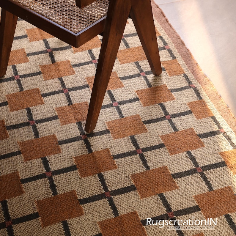 Op de afbeelding: Een geweven vloerkleed met een geometrisch patroon van bruine vierkanten en zwarte kruislijnen op een beige achtergrond. De houten poten van een stoel bedekken het kleed gedeeltelijk. Het kleed heeft een rand en de tekst "RugscreatioIN" is zichtbaar.