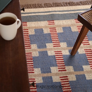 May include: A blue, beige, and red geometric patterned rug with a fringed edge. A white mug with tea sits on a wooden table. A wooden chair leg is visible on the right side of the rug. The rug has a repeating pattern.