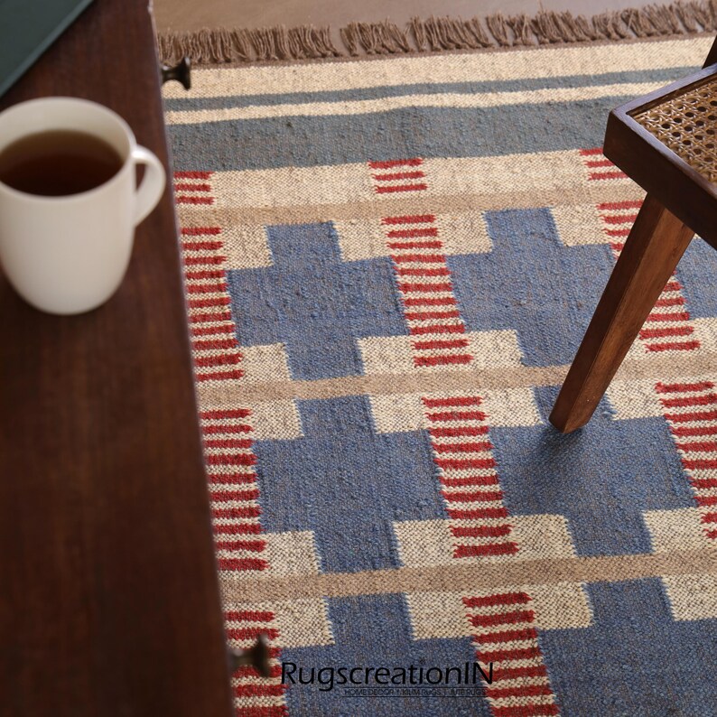 May include: A blue, beige, and red geometric patterned rug with a fringed edge. A white mug with tea sits on a wooden table. A wooden chair leg is visible on the right side of the rug. The rug has a repeating pattern.