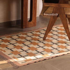 Op de afbeelding: Een rechthoekig tapijt met een geometrisch patroon van bruin, oranje en zwarte vierkanten. Het tapijt heeft een roze rand en een gefranjerde rand. Het tapijt ligt op een betegelde vloer, met een houten stoel- en tafelpoot zichtbaar.