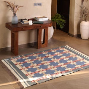 May include: A rectangular rug with a geometric pattern in blue, beige, and red. The rug has a gray border and fringe on the short ends. The rug is on a tiled floor in front of a wooden table.