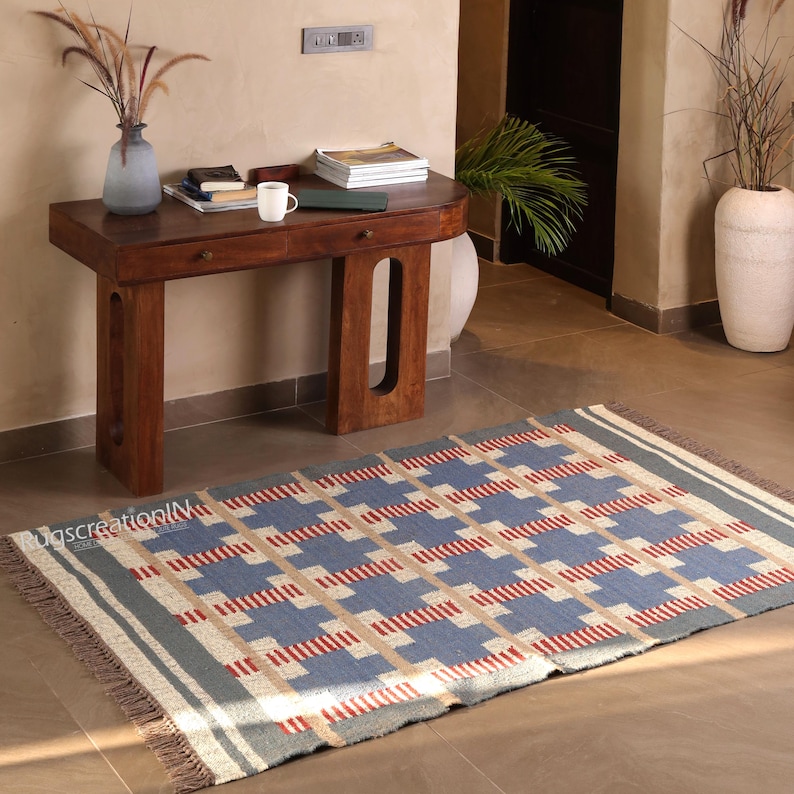 May include: A rectangular rug with a geometric pattern in blue, beige, and red. The rug has a gray border and fringe on the short ends. The rug is on a tiled floor in front of a wooden table.