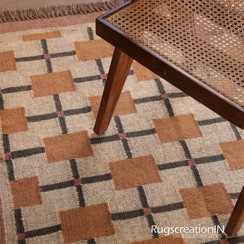 Op de afbeelding: Een beige tapijt met een geometrisch patroon van bruine vierkanten en zwarte kruisen. Een houten stoel met een geweven zitting is gedeeltelijk zichtbaar. Het tapijt heeft een gefranjerde rand en de tekst "RugscreatioN" is zichtbaar.
