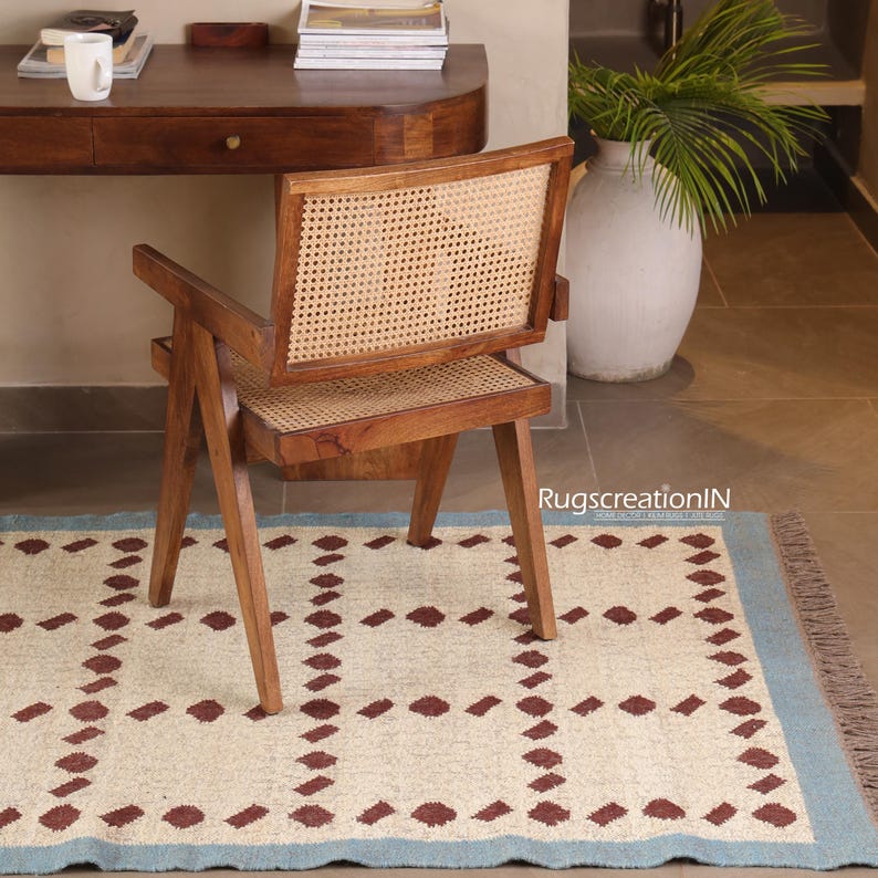 Puede incluir: Una silla de madera con respaldo y asiento de mimbre tejido se encuentra sobre una alfombra estampada. La silla tiene un acabado de madera natural y un respaldo rectangular. La alfombra tiene un fondo crema con un patr&oacute;n repetido de formas de diamante granate y un borde azul.