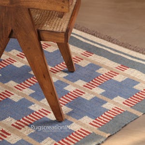 May include: A blue, beige, and red geometric patterned rug with a wooden chair leg in the foreground. The rug features a repeating pattern of blue squares, beige rectangles, and red stripes. The chair leg is made of wood.