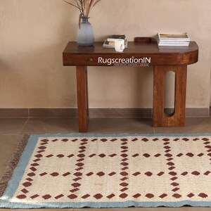 Puede incluir: Una alfombra rectangular con fondo crema y un patr&oacute;n repetitivo de formas de diamante granate. La alfombra tiene un borde azul claro y flecos. Una mesa de madera oscura con un jarr&oacute;n y libros est&aacute; en el fondo. El texto "RugscreatioIN" est&aacute; en la mesa.