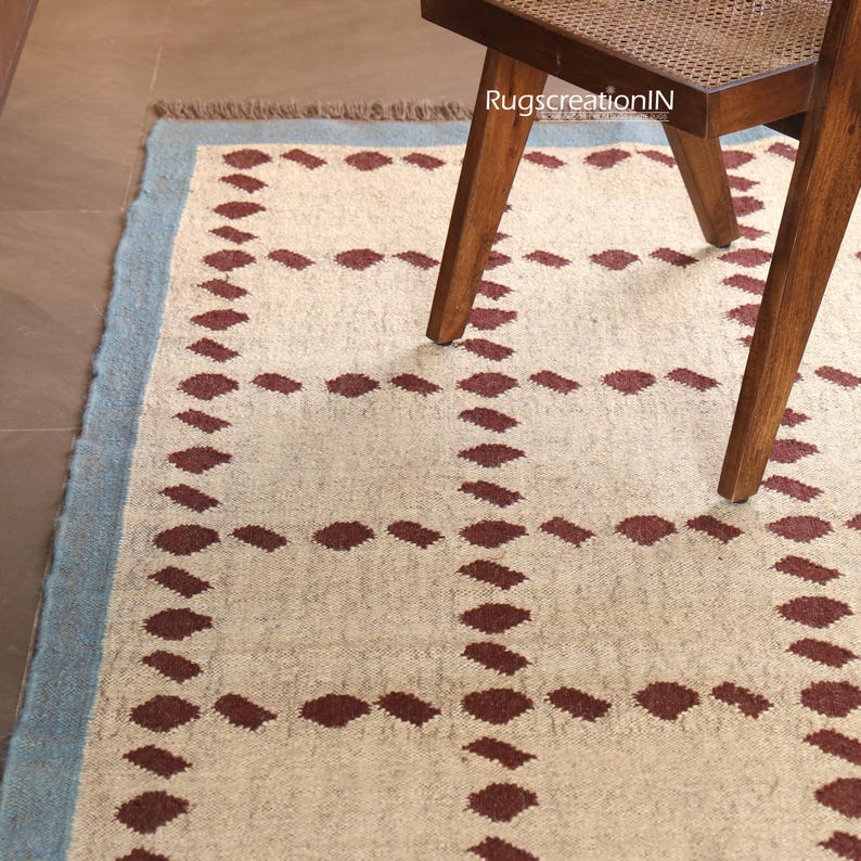 Puede incluir: Una alfombra rectangular con fondo crema y un patr&oacute;n repetitivo de rombos granate. La alfombra tiene un borde azul claro y est&aacute; parcialmente cubierta por una silla de madera.