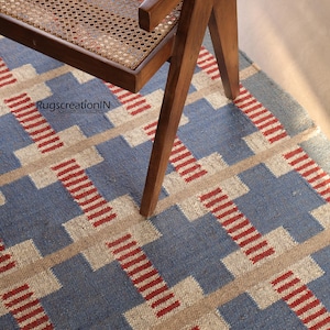 May include: A blue, beige, and red geometric patterned rug with a wooden chair leg and part of the chair visible. The rug has a repeating pattern of blue squares and beige rectangles with red stripes. The text "RugscreatioIN" is visible.