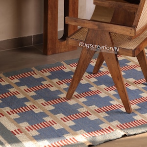 May include: A blue, beige, and red geometric patterned rug is partially visible beneath a wooden chair. The rug's design features a repeating pattern of rectangles and lines. The chair is made of wood and has a woven seat. The text "RugscreationIN" is visible on the chair.