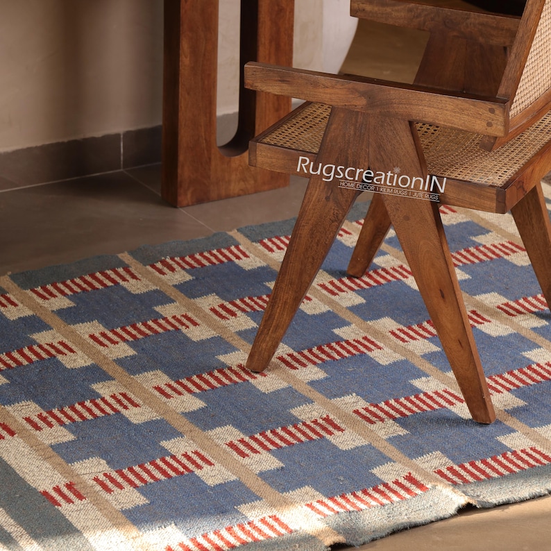 May include: A blue, beige, and red geometric patterned rug is partially visible beneath a wooden chair. The rug's design features a repeating pattern of rectangles and lines. The chair is made of wood and has a woven seat. The text "RugscreationIN" is visible on the chair.