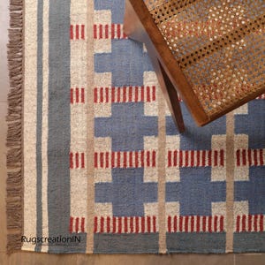 May include: A blue and beige geometric patterned rug with red accents and fringed edges. The rug has a woven texture and is partially covered by a wooden chair with a woven seat. The rug's design includes vertical and horizontal lines.