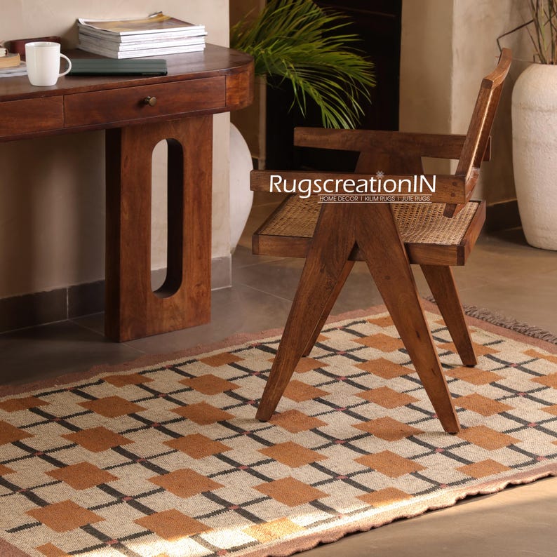 Op de afbeelding: Een geometrisch patroon tapijt met een beige achtergrond en bruine en zwarte vierkanten. Een houten stoel met een geweven zitting en rugleuning staat op het tapijt. De tekst "RugscreatioIN" is zichtbaar op de stoel. Een houten bureau en een witte mok staan op de achtergrond.