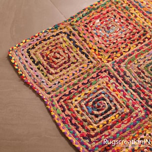 Puede incluir: Una colorida alfombra de yute hecha a mano con un diseño geométrico. La alfombra presenta un patrón repetitivo de cuadrados con centros en espiral, incorporando una variedad de colores como rojo, amarillo, verde y azul. Los bordes están cuidadosamente acabados con un borde trenzado.