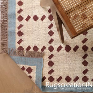Puede incluir: Alfombra beige y azul con un patr&oacute;n geom&eacute;trico de rombos granates. La alfombra tiene un borde azul y flecos en un borde. Una silla de madera con asiento tejido es parcialmente visible.