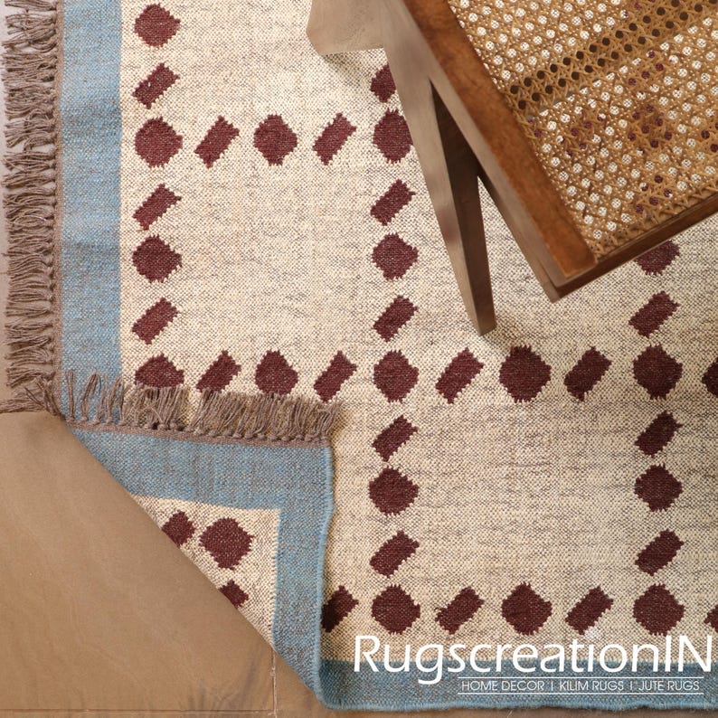 Puede incluir: Alfombra beige y azul con un patr&oacute;n geom&eacute;trico de rombos granates. La alfombra tiene un borde azul y flecos en un borde. Una silla de madera con asiento tejido es parcialmente visible.