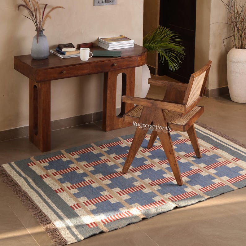 May include: A blue and beige geometric patterned rug with red accents, fringed edges, and a gray border. A wooden desk, chair, and decorative items are in the background, creating a warm, inviting interior design scene.