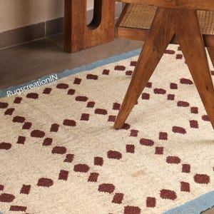 Puede incluir: Alfombra color crema con un patr&oacute;n repetitivo de cuadrados rojo oscuro. La alfombra tiene un borde azul claro y est&aacute; parcialmente oscurecida por una pata de mesa de madera. El texto "RugscreatioIN" es visible.