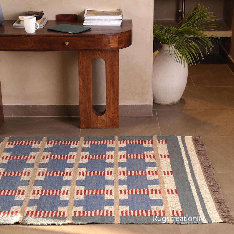 May include: A rectangular blue rug with a geometric pattern of red and beige rectangles. The rug has a fringed edge and a gray border. The rug is placed on a tiled floor near a wooden table and a white vase.