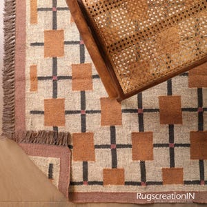 Op de afbeelding: Een geometrisch patroon tapijt met een bruine rand en franjes. Het tapijt heeft een herhalend patroon van bruine vierkanten en zwarte kruisvormen op een beige achtergrond. Een houten stoel met een geweven zitting is gedeeltelijk zichtbaar.