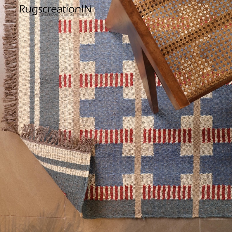 May include: A blue and beige geometric patterned rug with red accents and fringed edges. The rug has a woven texture and is partially covered by a wooden chair with a woven seat. The rug is labeled "RugscreatioIN".