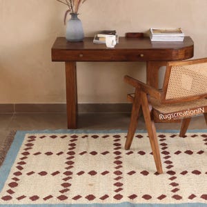 Puede incluir: Una alfombra crema con borde azul claro y un patr&oacute;n repetido de rombos granates. Un escritorio de madera, una silla y un jarr&oacute;n est&aacute;n en el fondo. La silla tiene respaldo y asiento de mimbre.