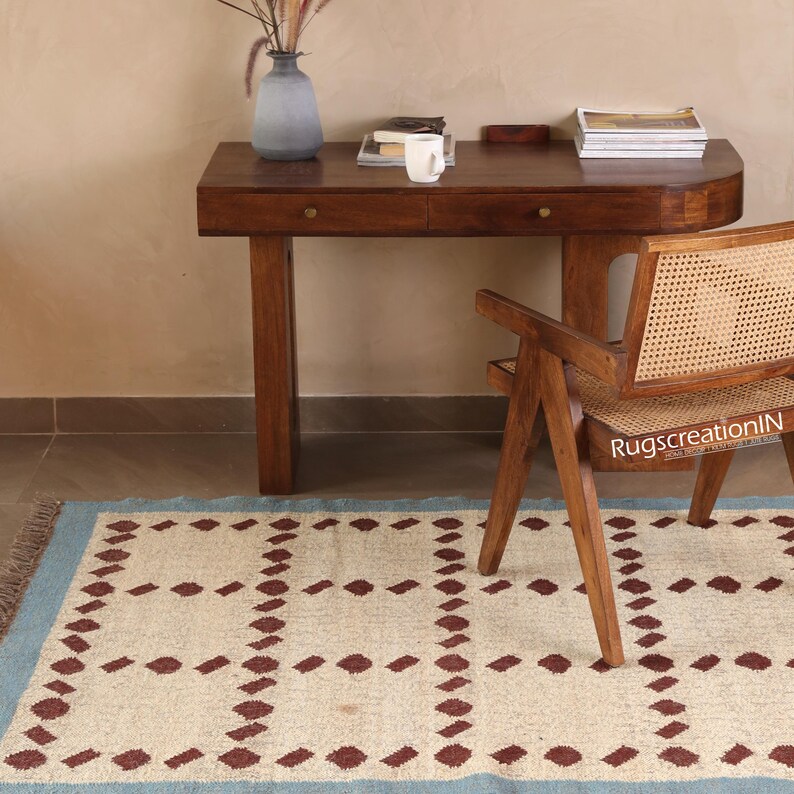 Puede incluir: Una alfombra crema con borde azul claro y un patr&oacute;n repetido de rombos granates. Un escritorio de madera, una silla y un jarr&oacute;n est&aacute;n en el fondo. La silla tiene respaldo y asiento de mimbre.