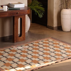 Op de afbeelding: Rechthoekig vloerkleed met een geometrisch patroon van beige, bruine en zwarte vierkanten. Het kleed heeft een bruine rand en franjes aan de uiteinden. Het ligt op een betegelde vloer voor een houten tafel.