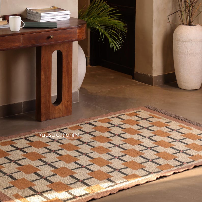 Op de afbeelding: Rechthoekig vloerkleed met een geometrisch patroon van beige, bruine en zwarte vierkanten. Het kleed heeft een bruine rand en franjes aan de uiteinden. Het ligt op een betegelde vloer voor een houten tafel.