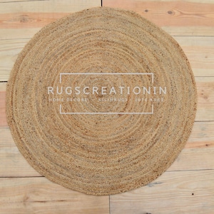 Puede incluir: Una alfombra redonda de yute con diseño en espiral, sobre un fondo de madera clara. La alfombra presenta el texto "RUGS CREATION IN" en un marco blanco. Las fibras naturales crean una superficie texturizada y neutra.