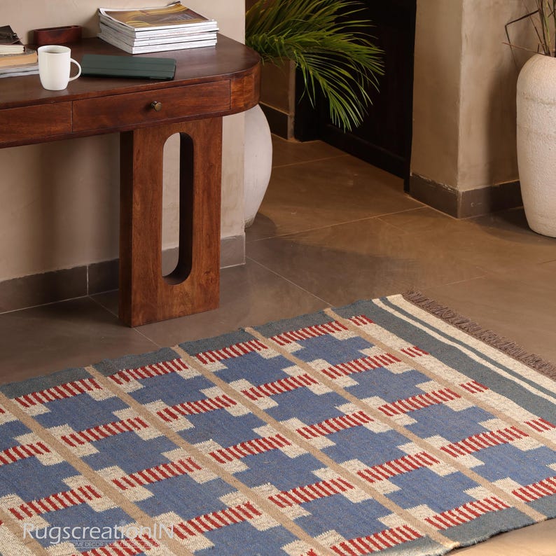 May include: A rectangular blue, red, and beige patterned rug with a geometric design. The rug has a fringe on one end and is placed on a tiled floor. The rug is in a room with a wooden table and a white vase.