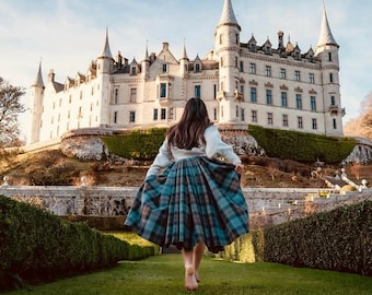 Può includere: Una persona di spalle indossa un maglione bianco e una gonna a quadri blu e marrone. Un grande castello con più guglie sullo sfondo, su una collina erbosa. Il cielo è limpido e blu.