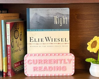 Może przedstawiać: Drewniany regał na książki prezentuje kilka książek, w tym "Night" Elie Wiesela, oraz znak z napisem "CURRENTLY READING". Szydełkowy słonecznik w białej doniczce dodaje dekoracyjnego akcentu. Półka wykonana jest z ciemnobrązowego drewna.