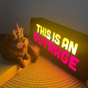 May include: A brown cat figurine wearing a crown and glasses rests on a book next to a black light-up sign. The sign displays the phrase "THIS IS AN OUTRAGE" in yellow and pink letters.