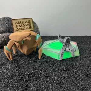 May include: A tan, multi-legged creature figurine with teal accents and a wooden sign reading "AMAZE" stands next to a grey, textured, seated figure perched on a hexagonal, glowing green light base, all set against a dark, granular surface.