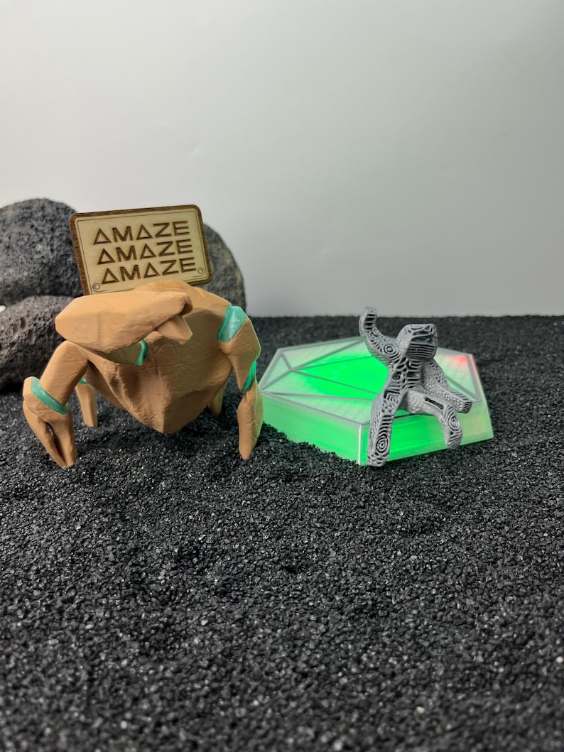 May include: A tan, multi-legged creature figurine with teal accents and a wooden sign reading "AMAZE" stands next to a grey, textured, seated figure perched on a hexagonal, glowing green light base, all set against a dark, granular surface.