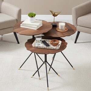 May include: Three wooden nesting tables with a distinctive scalloped edge, finished in a dark wood stain. The tables have black metal legs with gold-coloured tips. Accessories include a small plant, books, a candle, and a decorative bird. The tables are in a living room setting.