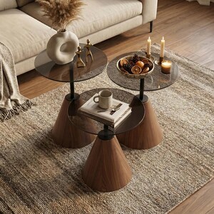 May include: A living room setting with a beige sofa, orange and cream cushions, and a patterned rug. Three side tables with dark wood conical bases and glass tops are arranged in front of the sofa. The tables hold decorative items like a vase, candles, and a bowl of dried fruit.