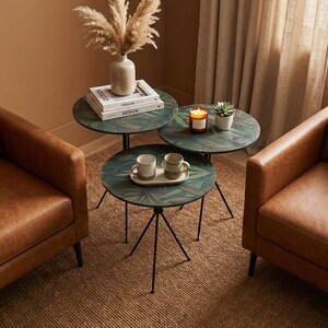 May include: Three round side tables with black metal legs and green leaf-patterned tops. The tables are clustered together and display books, a candle, and mugs. Two brown leather armchairs are positioned nearby.