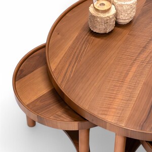 May include: A set of three wooden nesting tables with a warm brown finish. The tables have rounded tops and are supported by wooden legs. Two glass candle holders with decorative bases sit on the largest table.