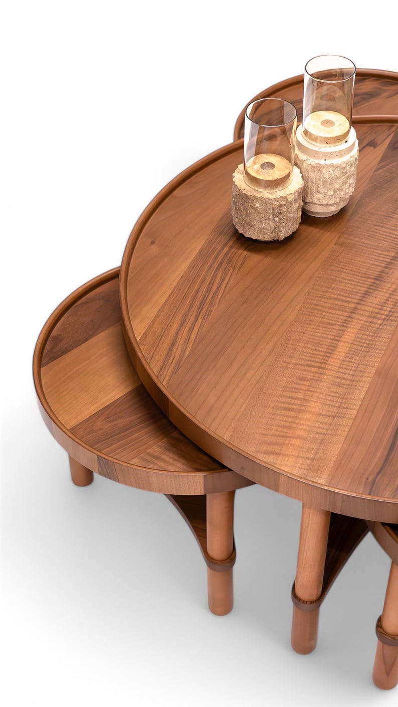 May include: A set of three wooden nesting tables with a warm brown finish. The tables have rounded tops and are supported by wooden legs. Two glass candle holders with decorative bases sit on the largest table.
