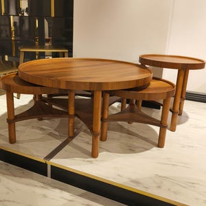 May include: A collection of four wooden coffee tables of different sizes. The tables have circular tops and light brown legs. The largest table is central, with smaller tables nested around it. The tables are made of wood.