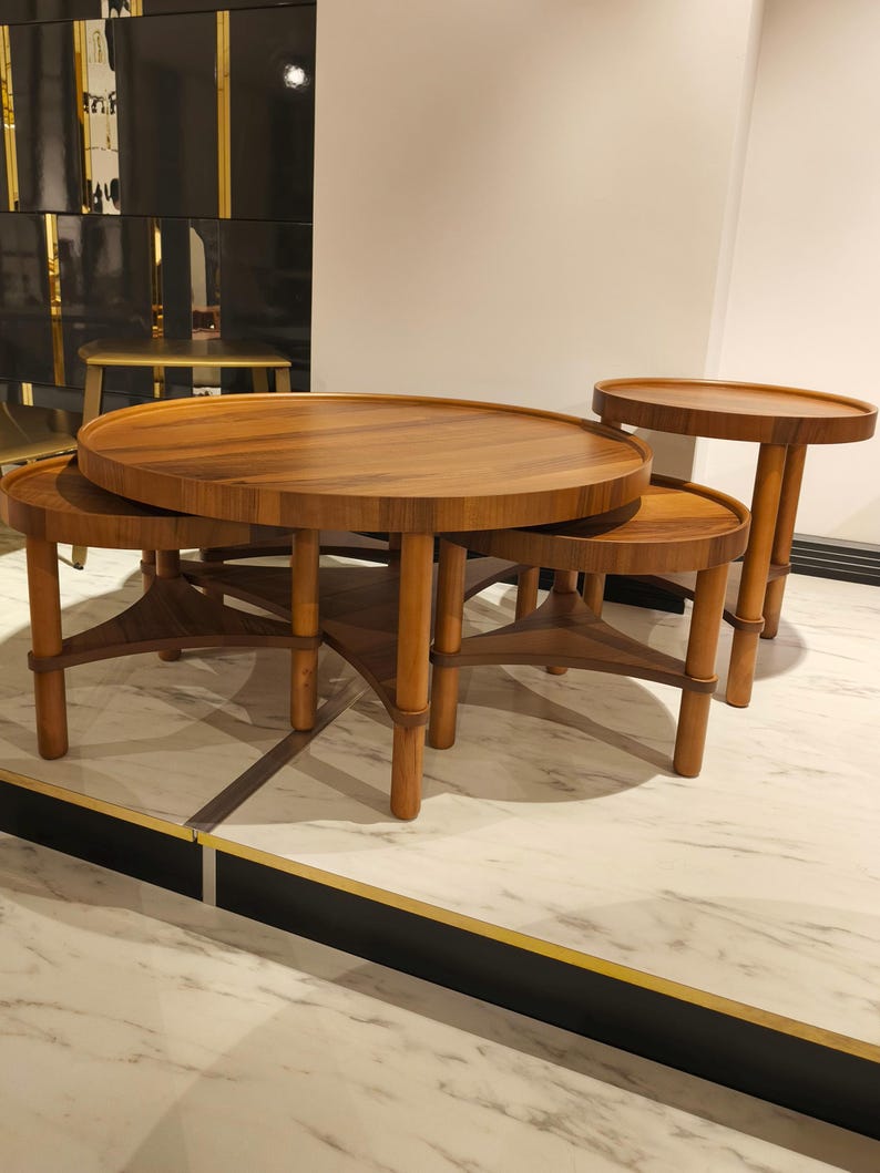 May include: A collection of four wooden coffee tables of different sizes. The tables have circular tops and light brown legs. The largest table is central, with smaller tables nested around it. The tables are made of wood.