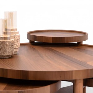 May include: A set of contemporary, circular wooden coffee tables in different sizes. The tables are crafted from dark brown wood with a smooth, polished finish. Two glass candle holders with a textured base are placed on the largest table.