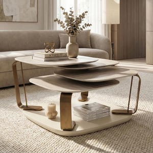 Op de afbeelding: Een moderne, gelaagde salontafel met een crèmekleurige basis en drie houten, bladvormige oppervlakken. De tafel heeft goudkleurige, gebogen metalen poten. Een stapel boeken met het woord "SCULPTURE" staat op het bovenste oppervlak.