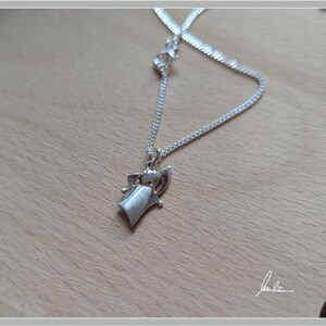 Peut inclure: Un collier avec pendentif argent&eacute; repr&eacute;sentant un ange stylis&eacute;. Le pendentif est suspendu &agrave; une cha&icirc;ne d&eacute;licate, pos&eacute; sur une surface en bois brun clair. L'ange a une robe fluide et des ailes d&eacute;ploy&eacute;es.